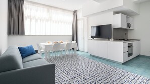 Un apartamento moderno con un sofá gris, una mesa de comedor con sillas, una cocina con armarios blancos y un ventanal con cortinas translúcidas.