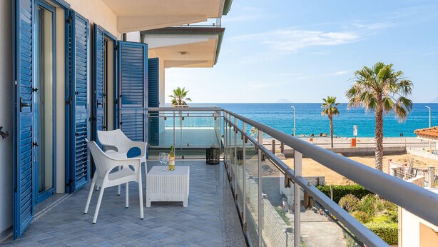 Un balcón con una silla y una mesa blancas, una botella de vino y vistas al mar y palmeras.