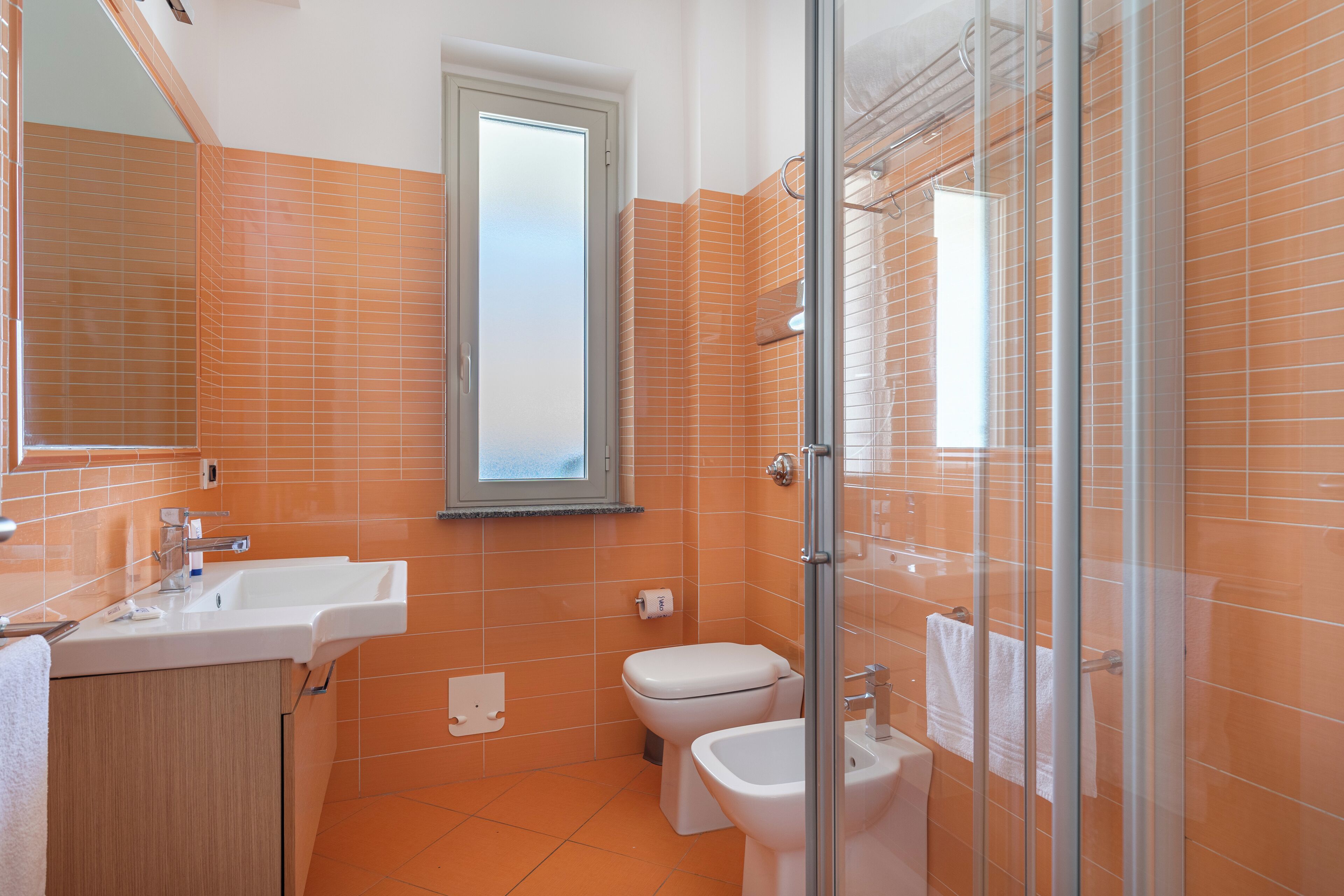 A bathroom with orange tiles, a white sink, a toilet, and a glass shower enclosure.