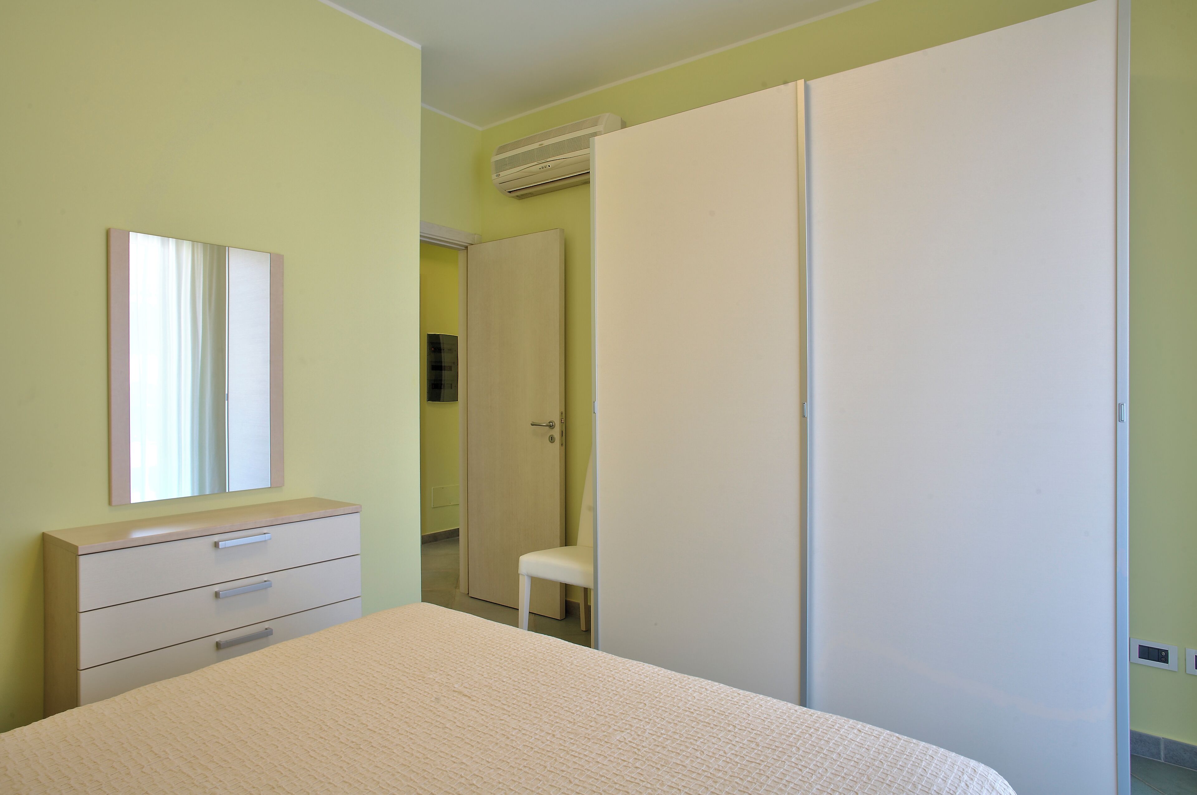 A bedroom with a bed, a dresser, a mirror, a wardrobe, and an air conditioning unit.