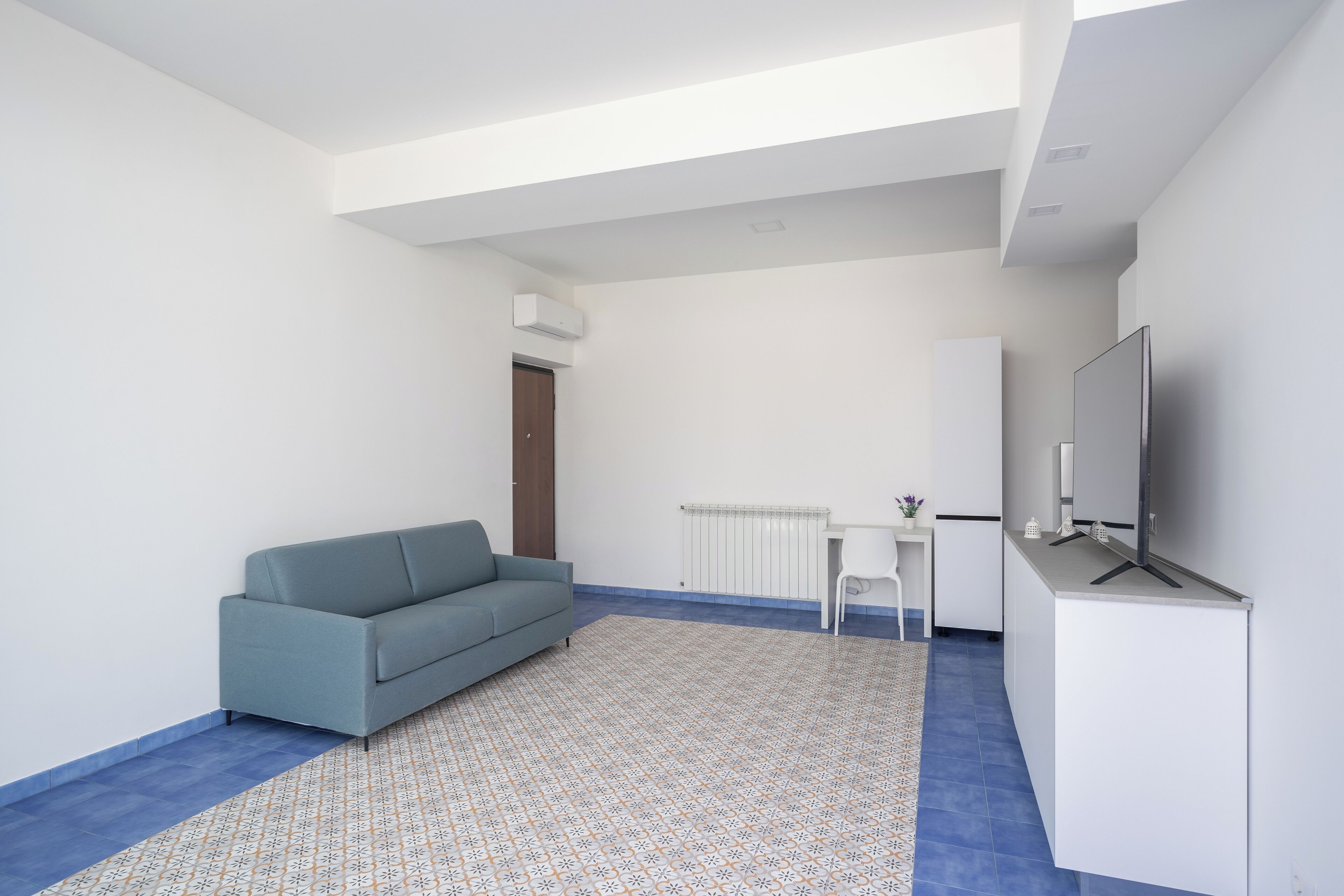 Een moderne, minimalistische woonkamer met een grijze bank, een klein wit tafeltje, een flatscreen televisie en een kamerkleed met een patroon.