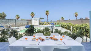 Un área de comedor al aire libre con una mesa dispuesta para dos personas, rodeada de sillas y vistas a una piscina y palmeras.