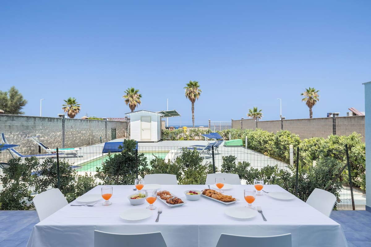 Un espace de restauration en plein air avec une table dressée pour deux personnes, entourée de chaises, et offrant une vue sur une piscine et des palmiers.