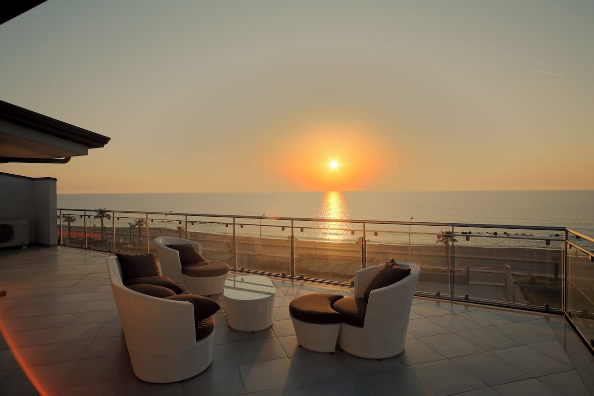 Un balcon doté de meubles en osier et d’une rambarde en verre, offrant une vue sur la mer au coucher du soleil.
