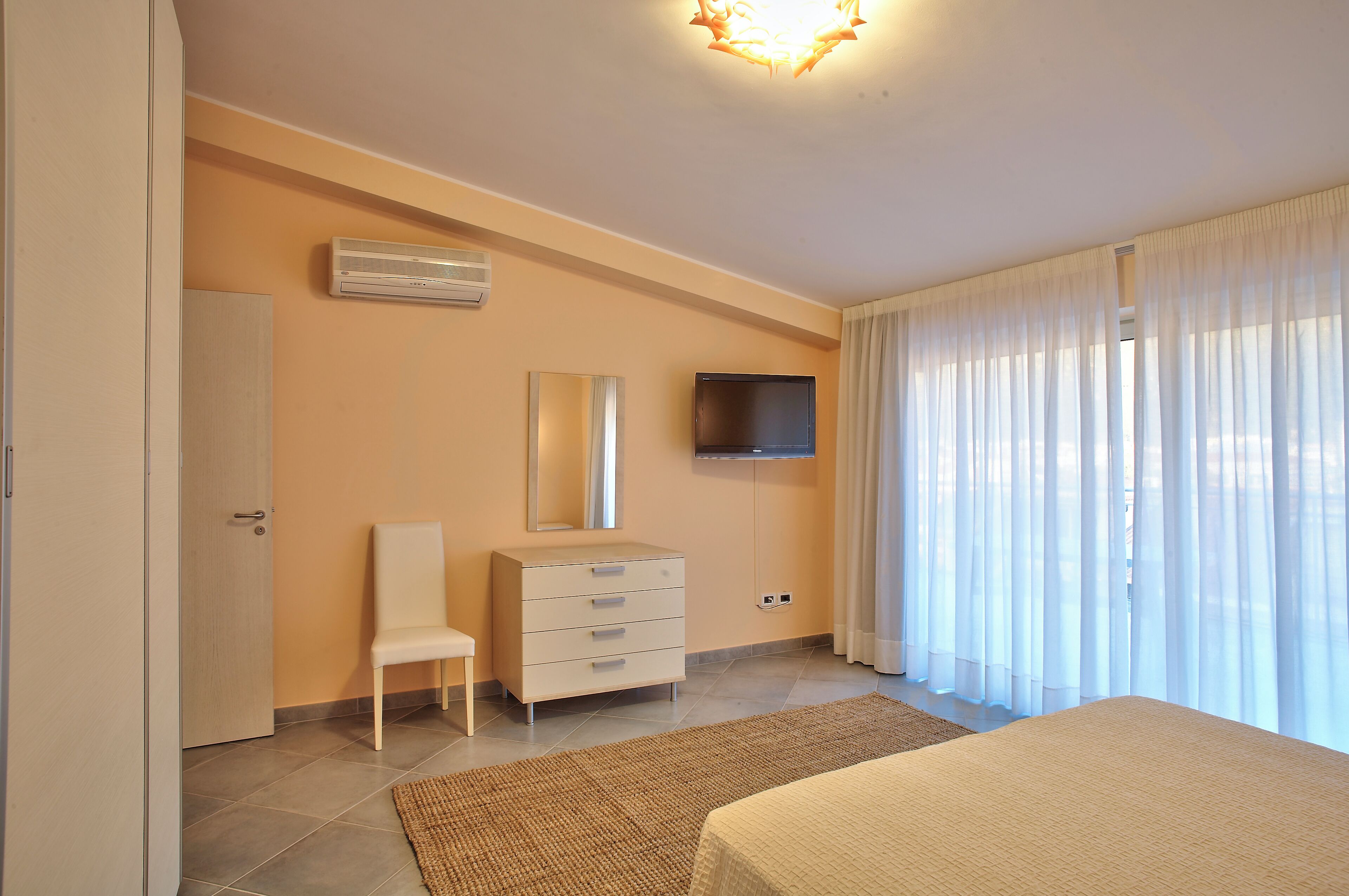 A bedroom with a bed, a chair, a dresser, a TV, and a window with curtains.