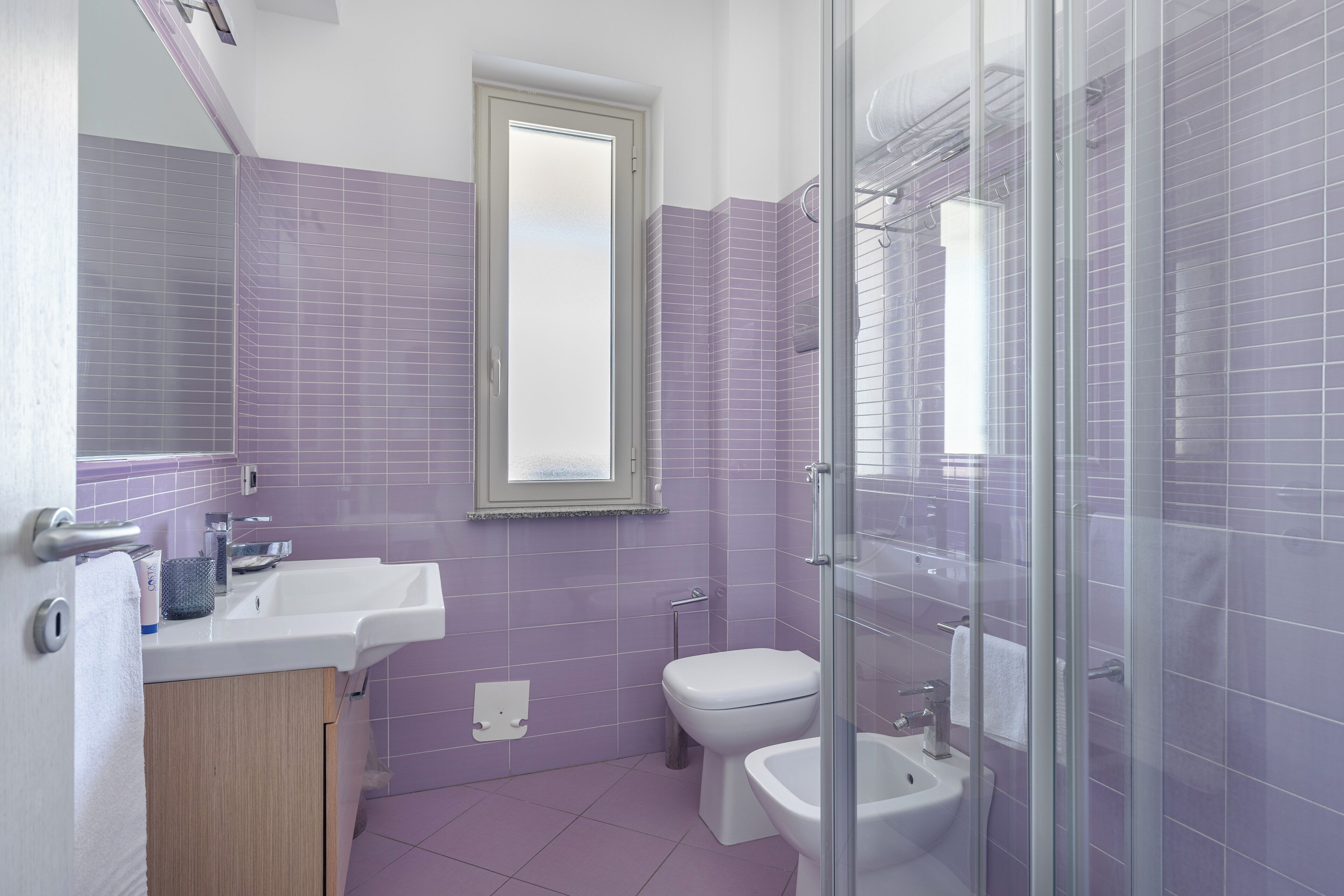 A bathroom with a sink, toilet, and bidet, featuring purple tiles and a glass shower enclosure.