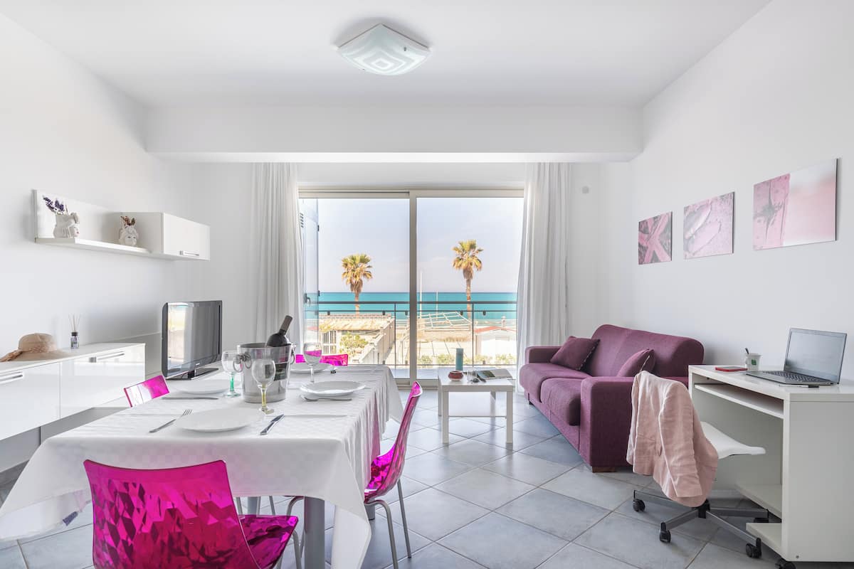 Un salon moderne comprenant un coin repas, un canapé violet et un balcon offrant une vue sur les palmiers et la mer.