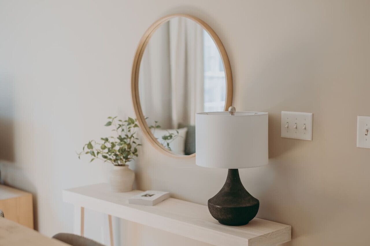 A minimalist room with a round mirror, a bedside table, a lamp, and a potted plant.
