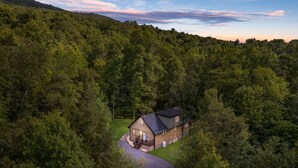 A house surrounded by dense forest, with a paved driveway leading to the entrance.