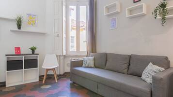 A living room with a grey sofa, a white chair, a small white shelf with plants, and a window with curtains.