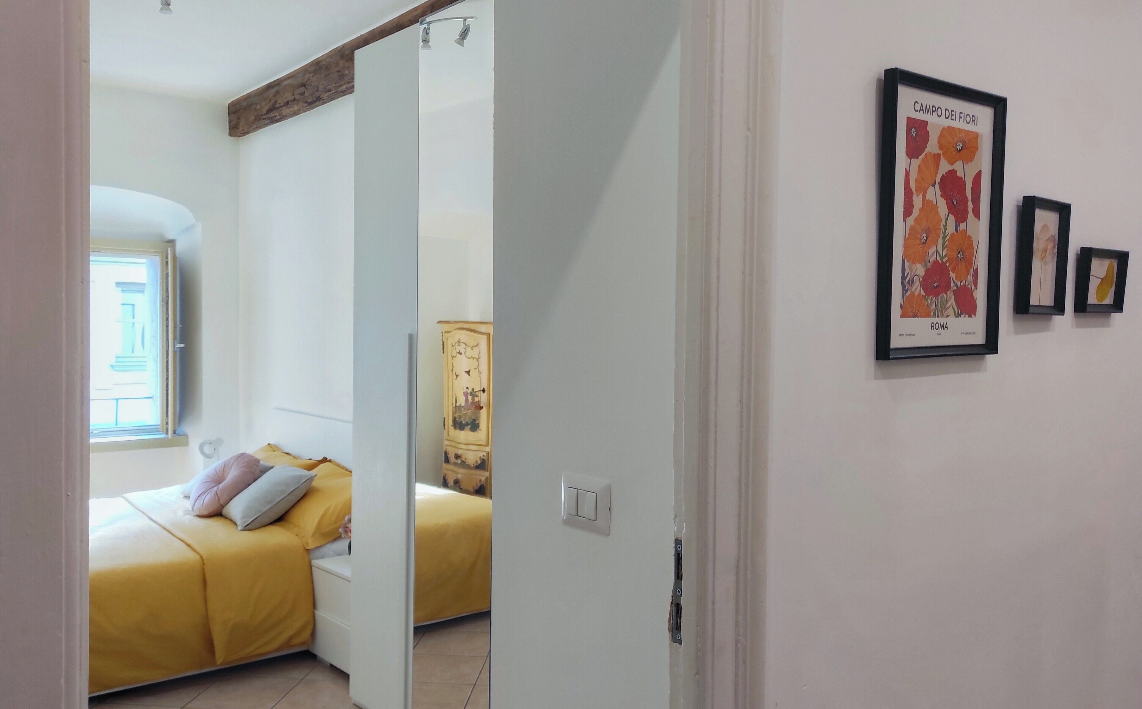 A bedroom with a bed, a window, and a door leading to another room. There are framed pictures on the wall.
