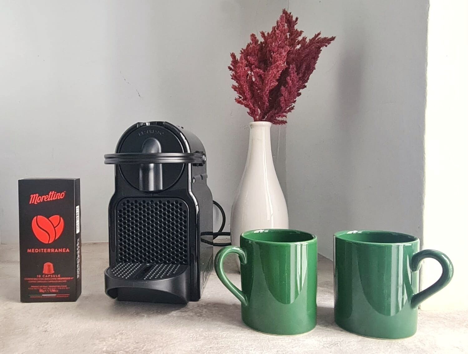 A coffee maker, a box of Morettino coffee capsules, a white vase with red flowers, and two green mugs.