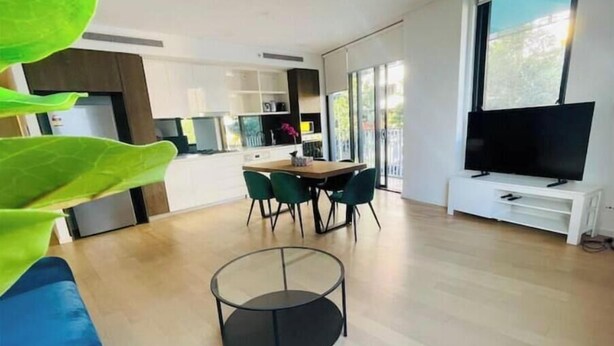 A modern living room with a round glass coffee table, a dining area with a wooden table and green chairs, a flat-screen TV on a white stand, and a kitchen in the background. - Amazing 2 bedrooms condo [Street Parking, WiFi] (Zetland)