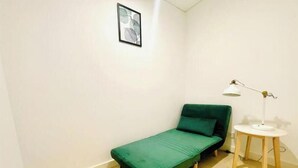 Ein kleines Zimmer mit einem Einzelbett, einer grünen Matratze, einem kleinen Nachttisch mit Lampe und einem gerahmten Bild an der Wand.