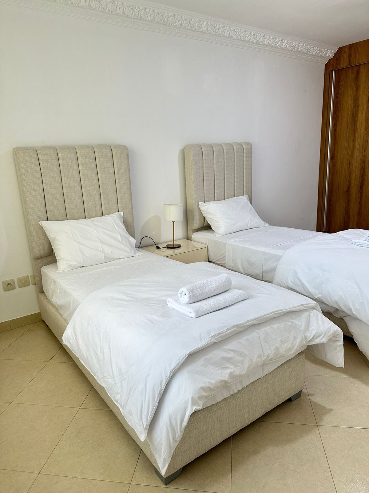 A hotel room with two beds, each with a headboard, a bedside table with a lamp, and a neatly made bed with a rolled towel.