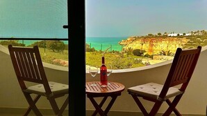 Un balcón con dos sillas de madera, una mesita redonda y una botella de vino. La vista muestra un paisaje costero con acantilados y cielo despejado.