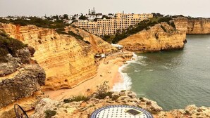 Vista costera con playa rocosa, mesa y sillas al borde de un acantilado, y un edificio en el fondo.