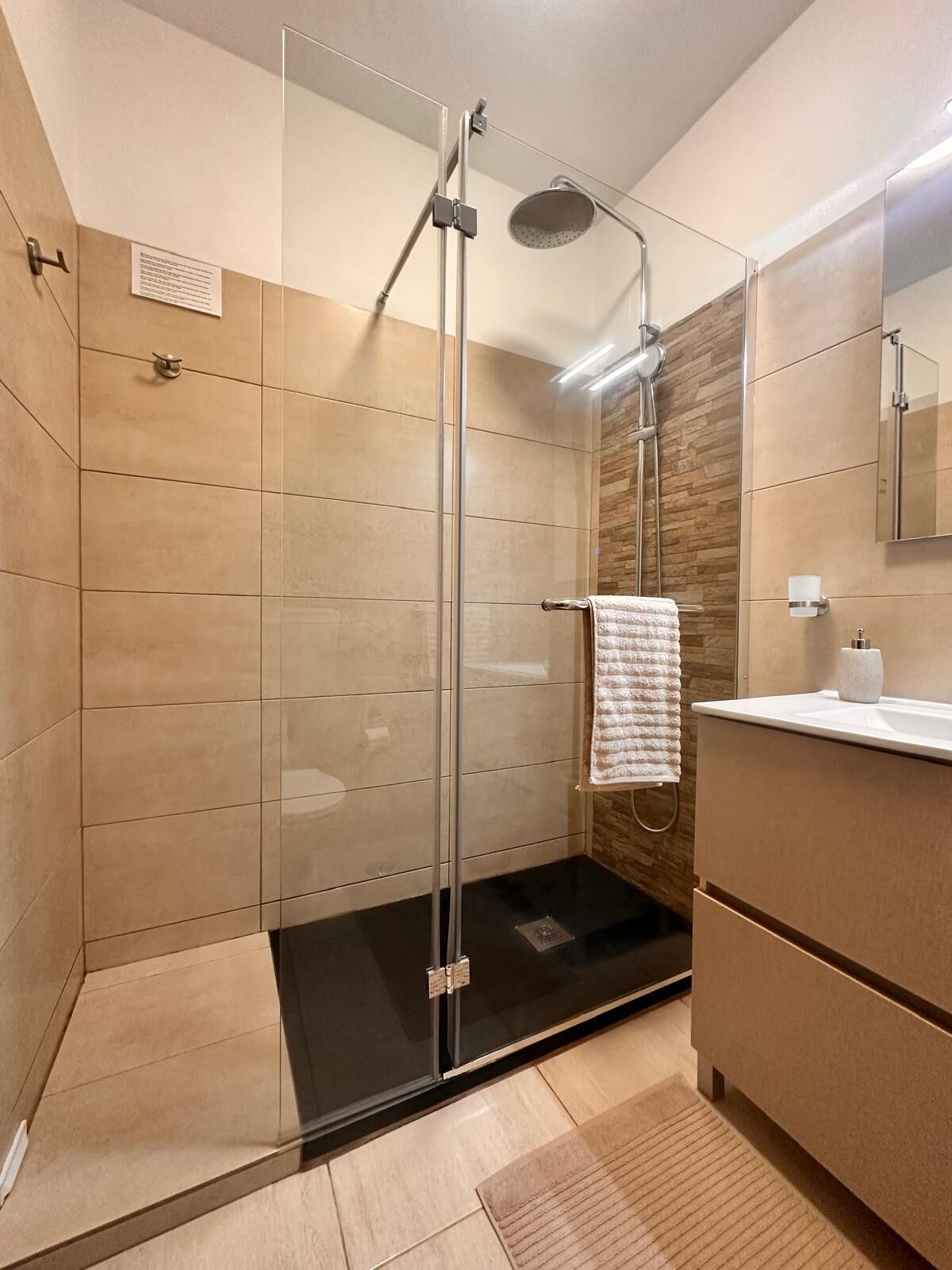 A modern bathroom with a glass-enclosed shower, a wooden wall, a white sink, and a mirror.