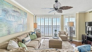 A living room with a large painting, a ceiling fan, a sofa set, a television, and a view through large windows.