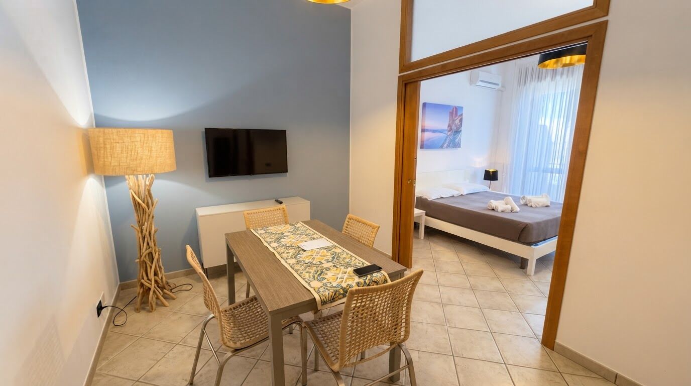 Habitación de hotel con cama, televisor montado en la pared, mesa de comedor con sillas y vista al mar a través de una ventana.