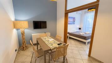 Habitación de hotel con cama, televisor montado en la pared, mesa de comedor con sillas y vista al mar a través de una ventana.