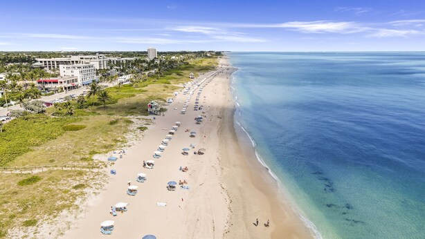 Ein Strand mit Liegestühlen und Sonnenschirmen, ein klares blaues Meer und Gebäude im Hintergrund.