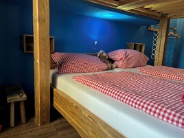 Una cama de madera con ropa de cama a cuadros rojos y blancos, un taburete de madera y una pared azul.