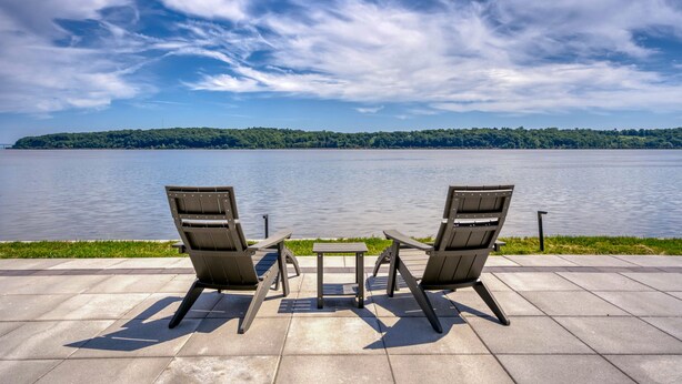 Two Adirondack chairs facing a lake with a small table in between, on a paved deck with a grassy area in the foreground and a forested hill in the background.