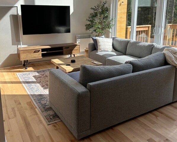 A modern living room with a grey sectional sofa, a flat-screen TV, a wooden coffee table, and a large window with a view of greenery. - Luxury chalet with fireplace, games room, close to popular ski resorts (Sainte-Lucie-des-Laurentides)
