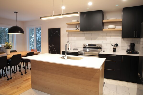 A modern kitchen with a central island, stainless steel appliances, black cabinets, and a pendant light. - Luxury chalet with fireplace, games room, close to popular ski resorts (Sainte-Lucie-des-Laurentides)