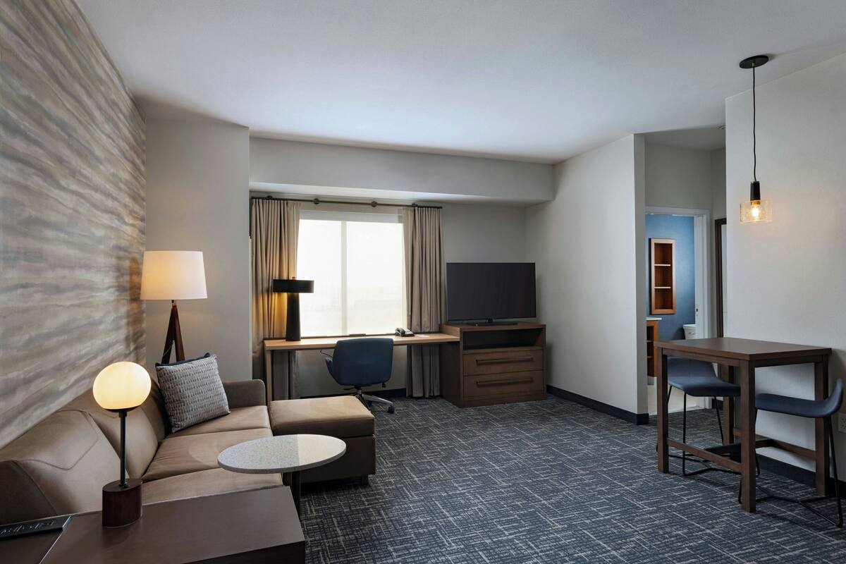 Habitación de hotel con sofá, una pequeña mesa redonda, escritorio con silla, televisor y lámpara.