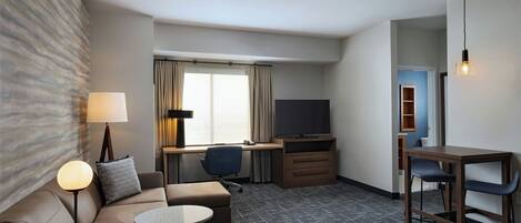 A hotel room with a sofa, a small round table, a desk with a chair, a television, and a lamp.