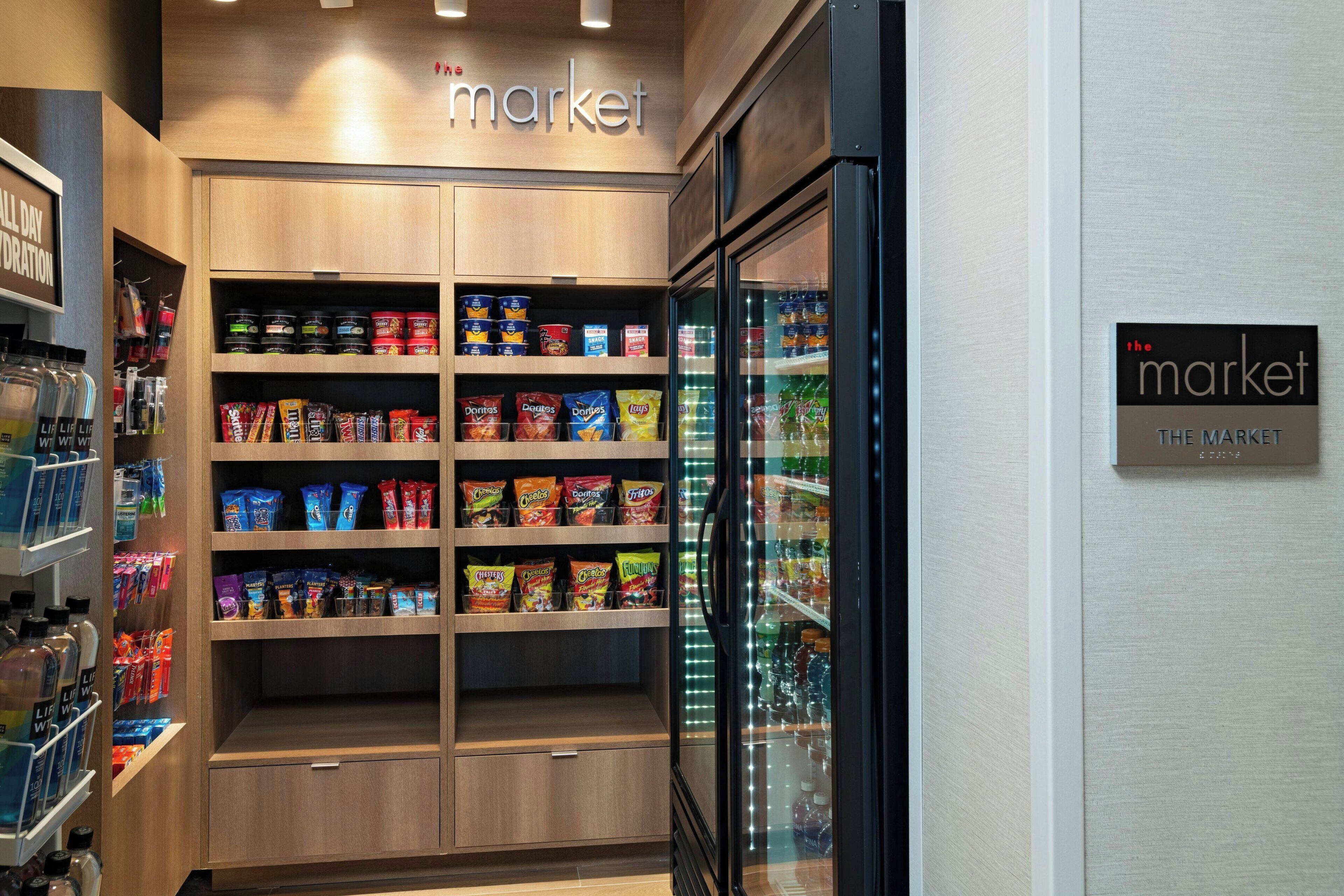 Un sector de una tienda con estantes llenos de diversas botanas y bebidas, un mostrador refrigerado y un letrero que indica el nombre de la tienda.