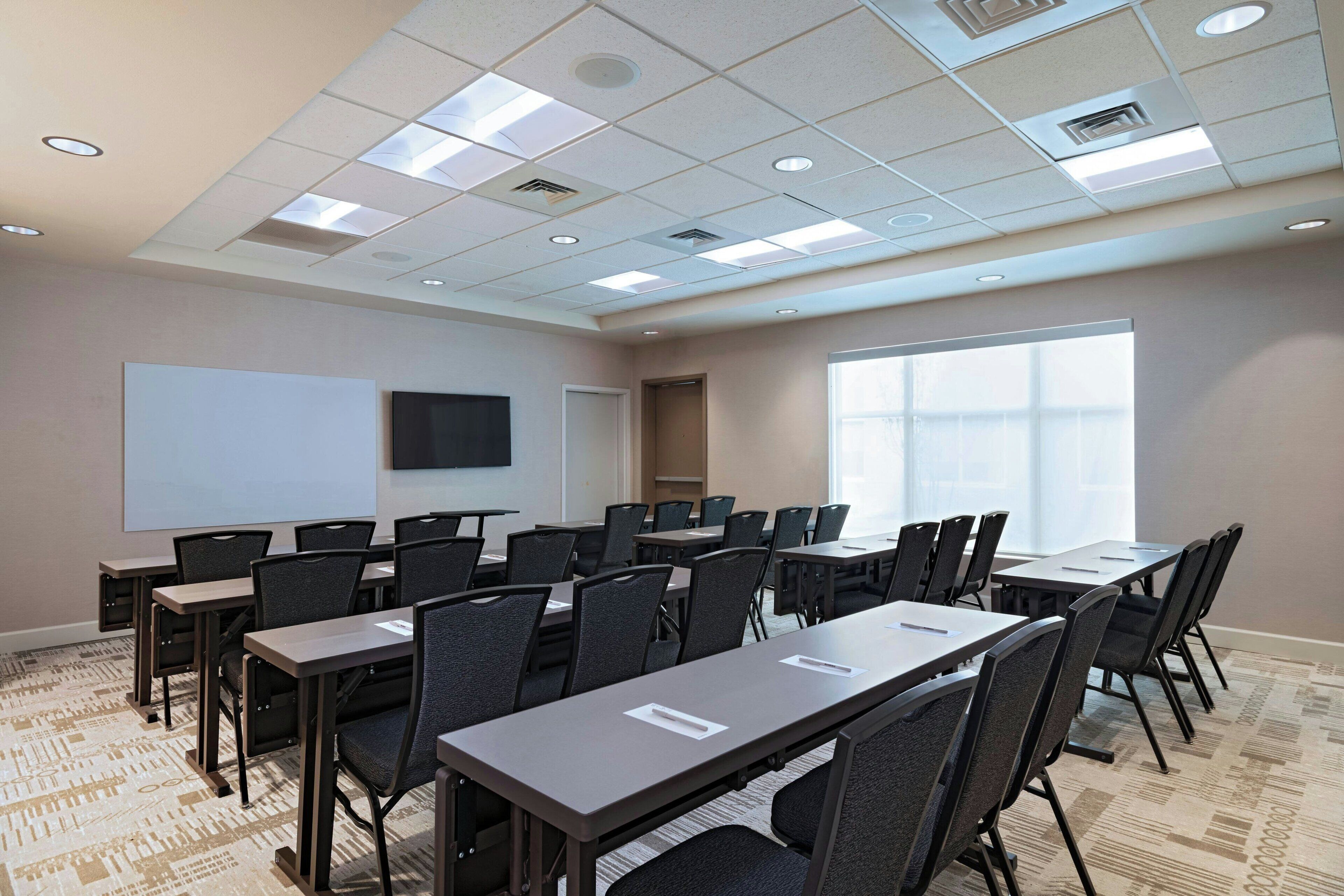 A conference room with multiple tables and chairs, a projector screen, and a large window providing natural light.