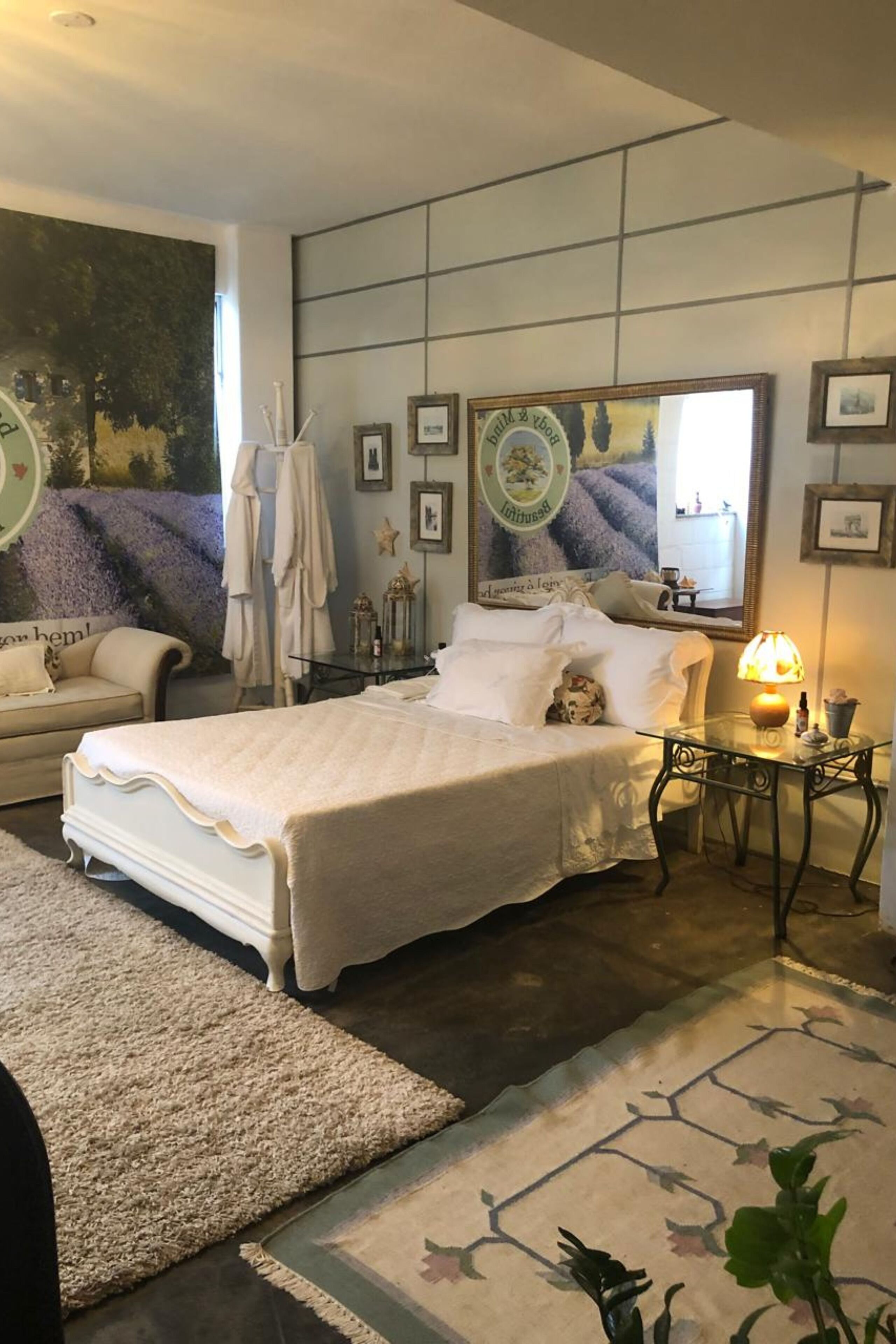 A bedroom with a large bed, a bedside table with a lamp, a mirror, and a wall with framed pictures.