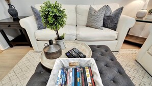 Een woonkamer met een witte bank, grijze kussens, een kleine potplant, een bijzettafeltje en een mand vol dvd's.