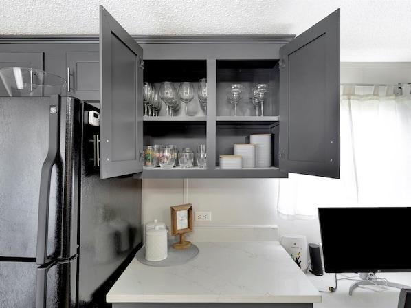 A kitchen with a black refrigerator, a white countertop, and a cabinet with wine glasses and plates.