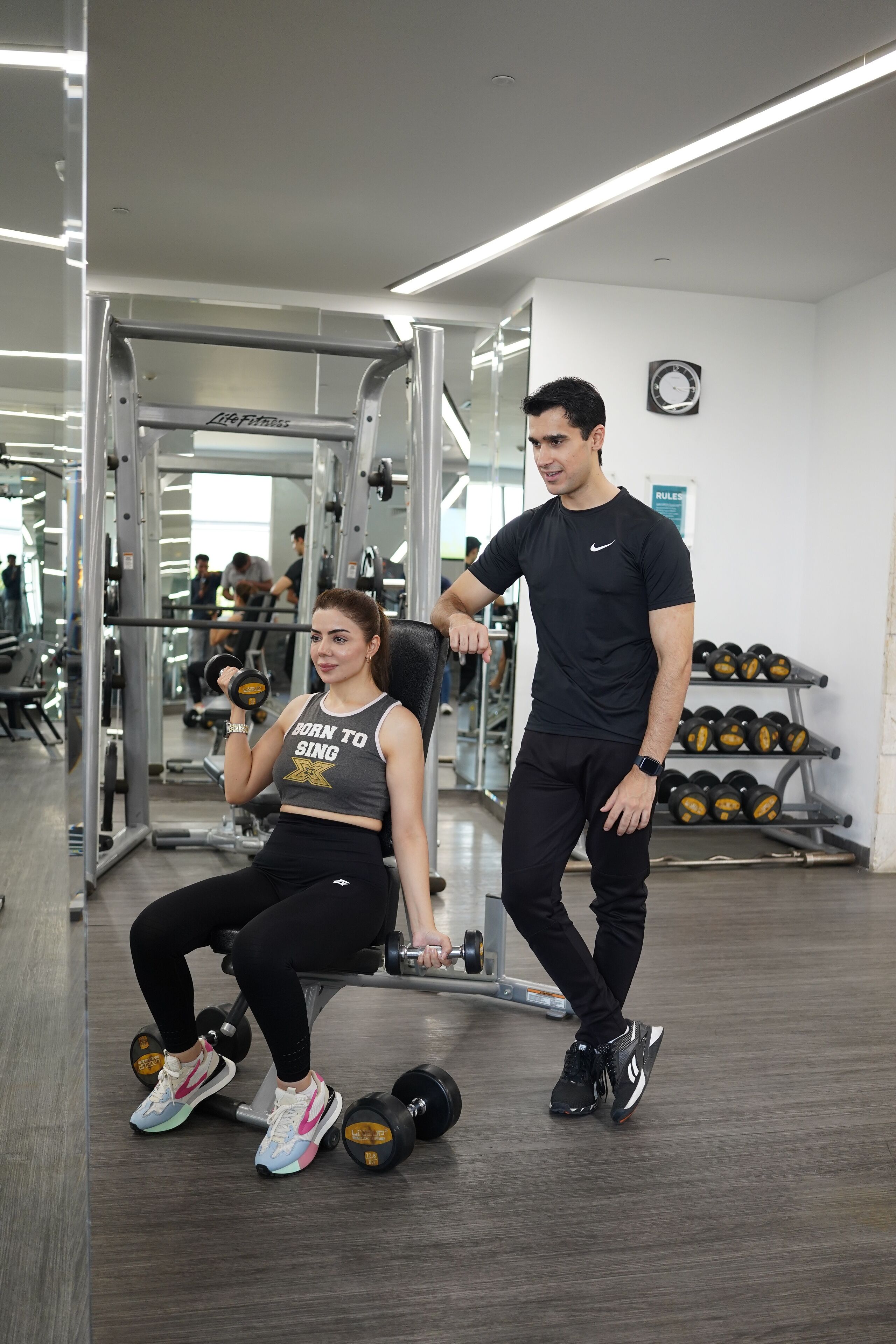 Ein Fitnessraum mit Trainingsgeräten, eine sitzende Frau, die Kurzhanteln benutzt, und ein stehender Mann, der zusieht.