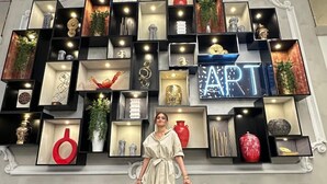 Une femme se tient devant un mur orné de plusieurs étagères où sont exposés divers objets décoratifs, et un néon affiche le mot « ART ».