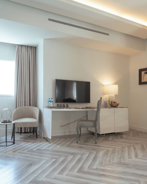 A hotel room with a flat-screen TV, a desk, a chair, a lamp, and a herringbone-patterned floor.