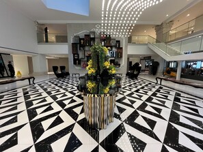 Un vestíbulo con piso de baldosas blanco y negro geométricas, una jardinera decorativa central con flores amarillas, y una escalera al fondo.