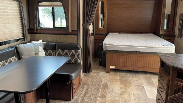 A compact RV interior with a bed, a dining area, and a seating space.