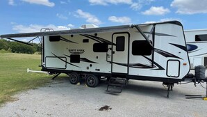 A White Hawk RV parked on a gravel surface with a grassy field in the background.