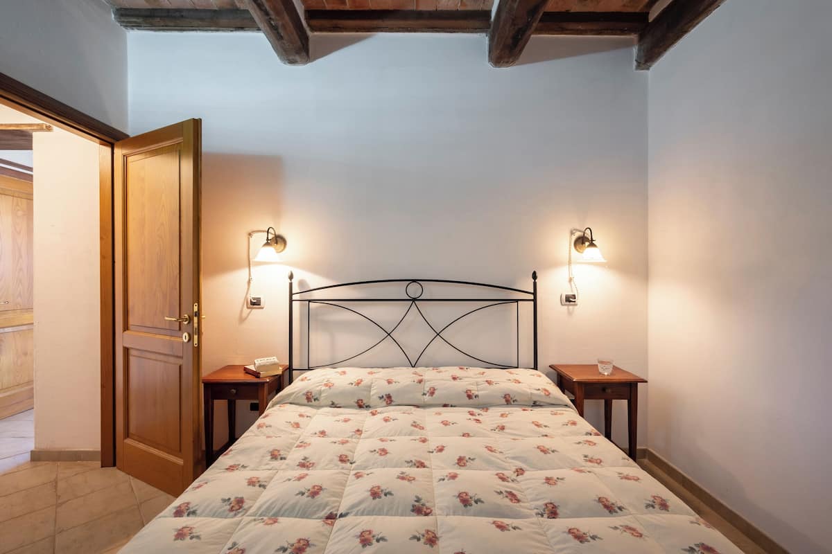 Un dormitorio con puerta de madera, una cama con ropa de cama floral, dos mesitas de noche con lámparas y un techo de madera.