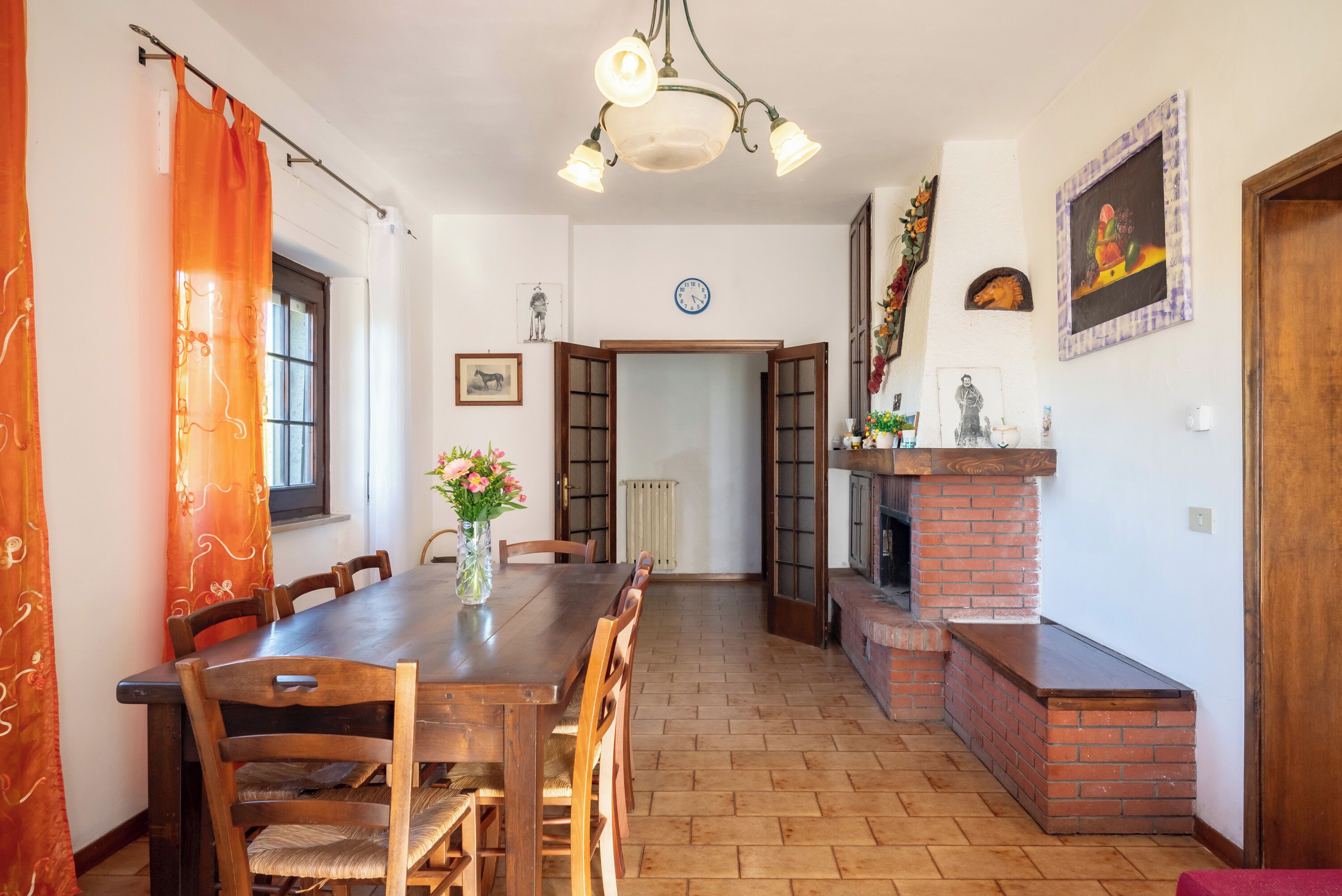 Uma sala de jantar com mesa e cadeiras de madeira, lareira de tijolos e um relógio na parede.