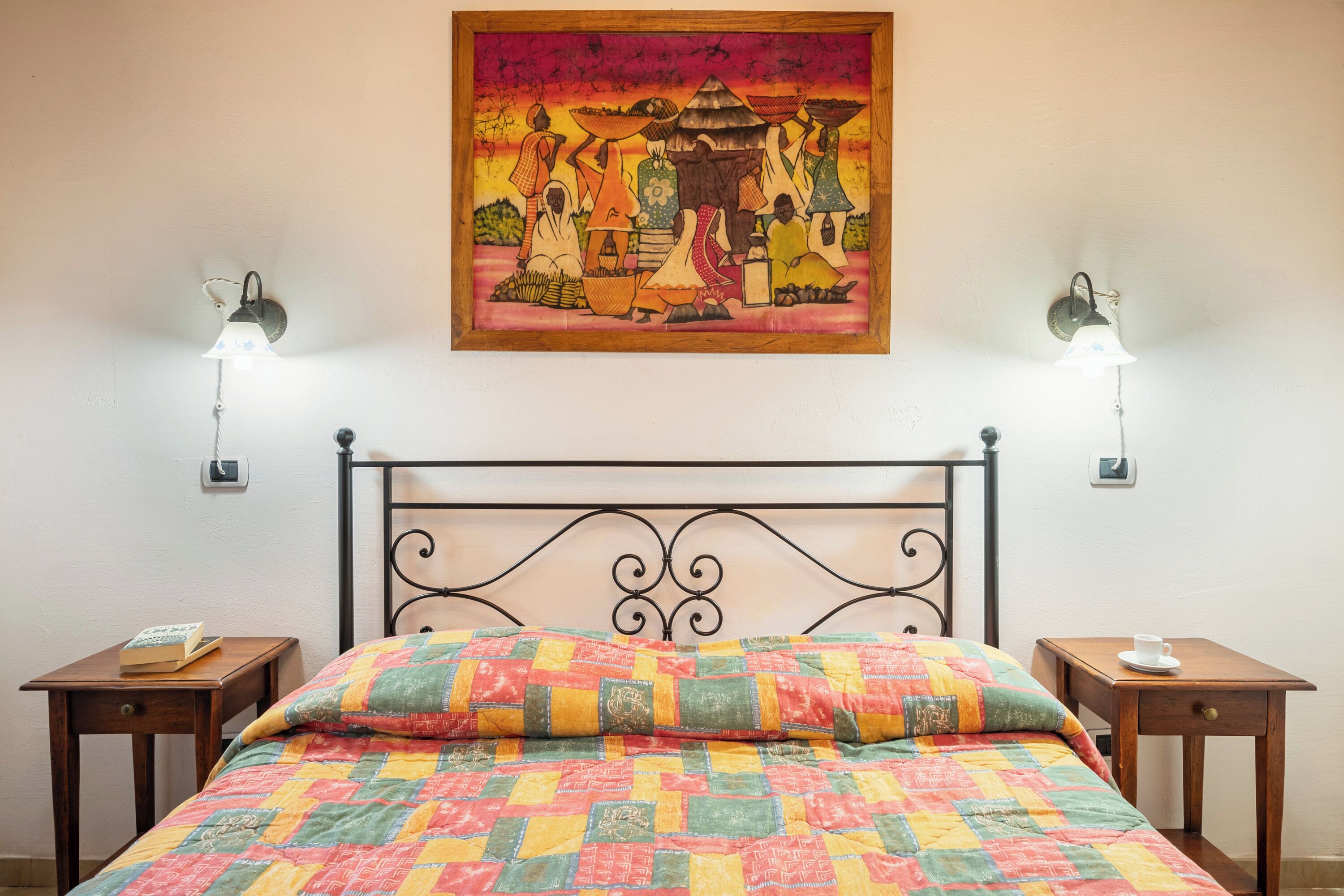 A bed with a colorful quilt, a painting above it, and two bedside tables with lamps.