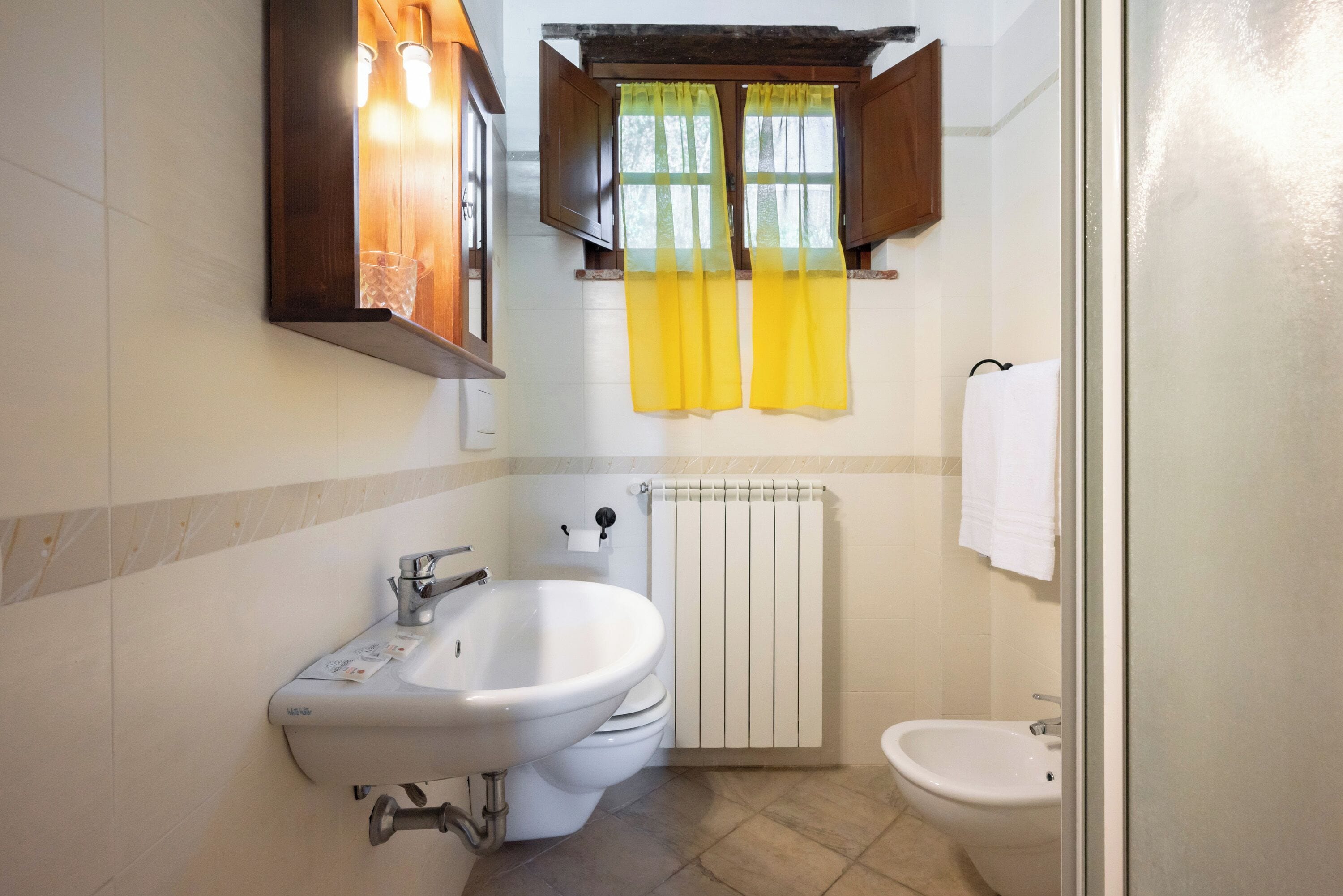 Un baño con lavamanos, inodoro y bidé, una ventana con cortinas amarillas y una toalla colgada en la pared.