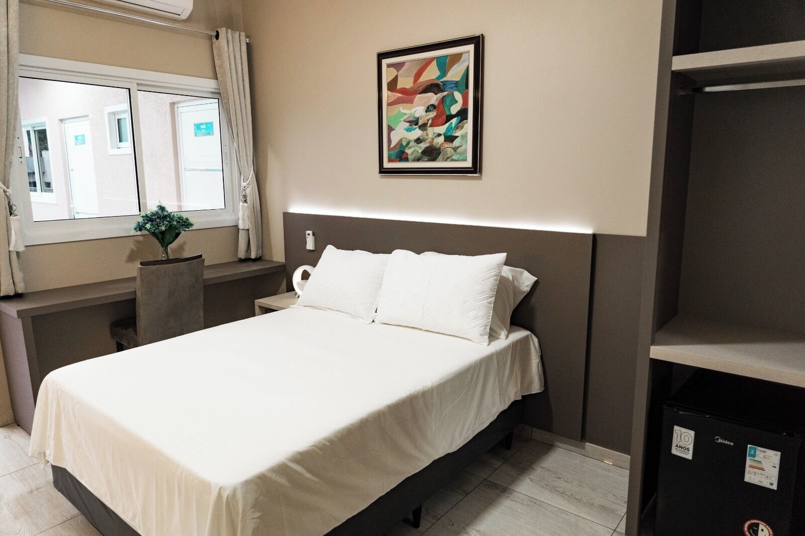 A hotel room with a bed, a nightstand, a wall-mounted painting, a window with curtains, and a potted plant.