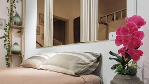Un lit bien fait, recouvert d’une courtepointe beige, une table de chevet en bois ornée d’un vase de fleurs roses et un miroir qui reflète la pièce.