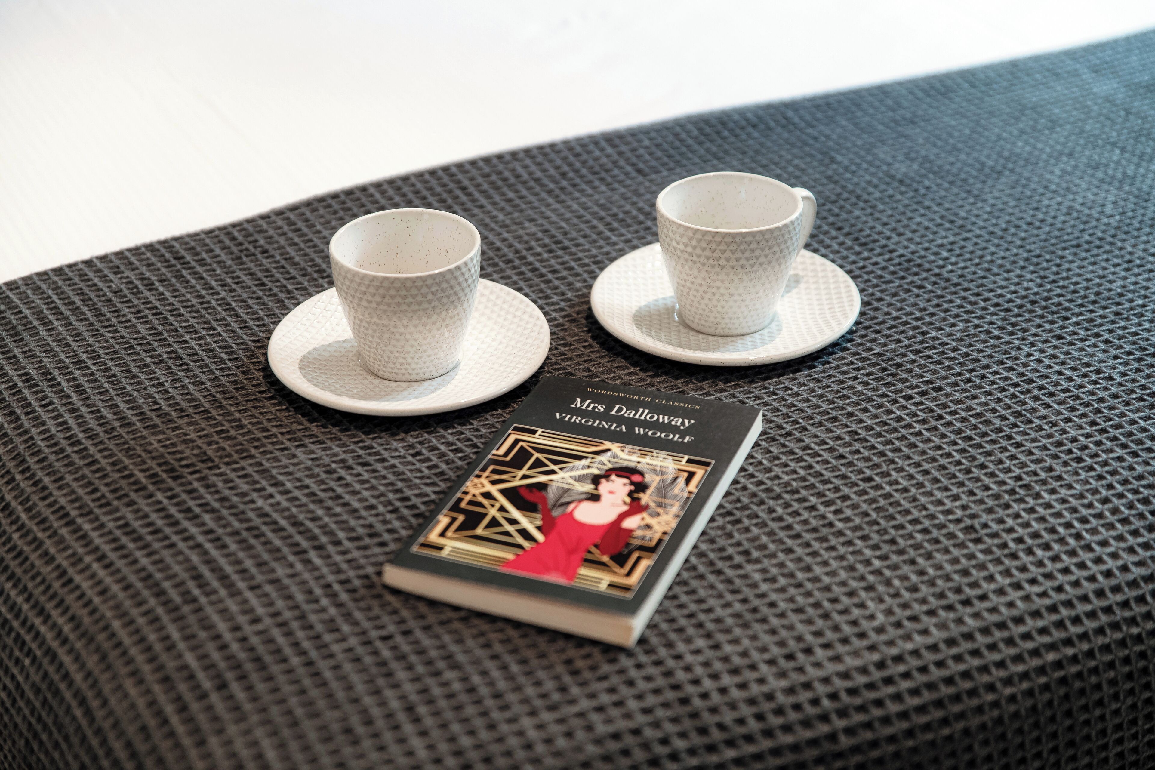 Two white cups on saucers and a book titled 'Mrs Dalloway' by Virginia Woolf on a textured surface.